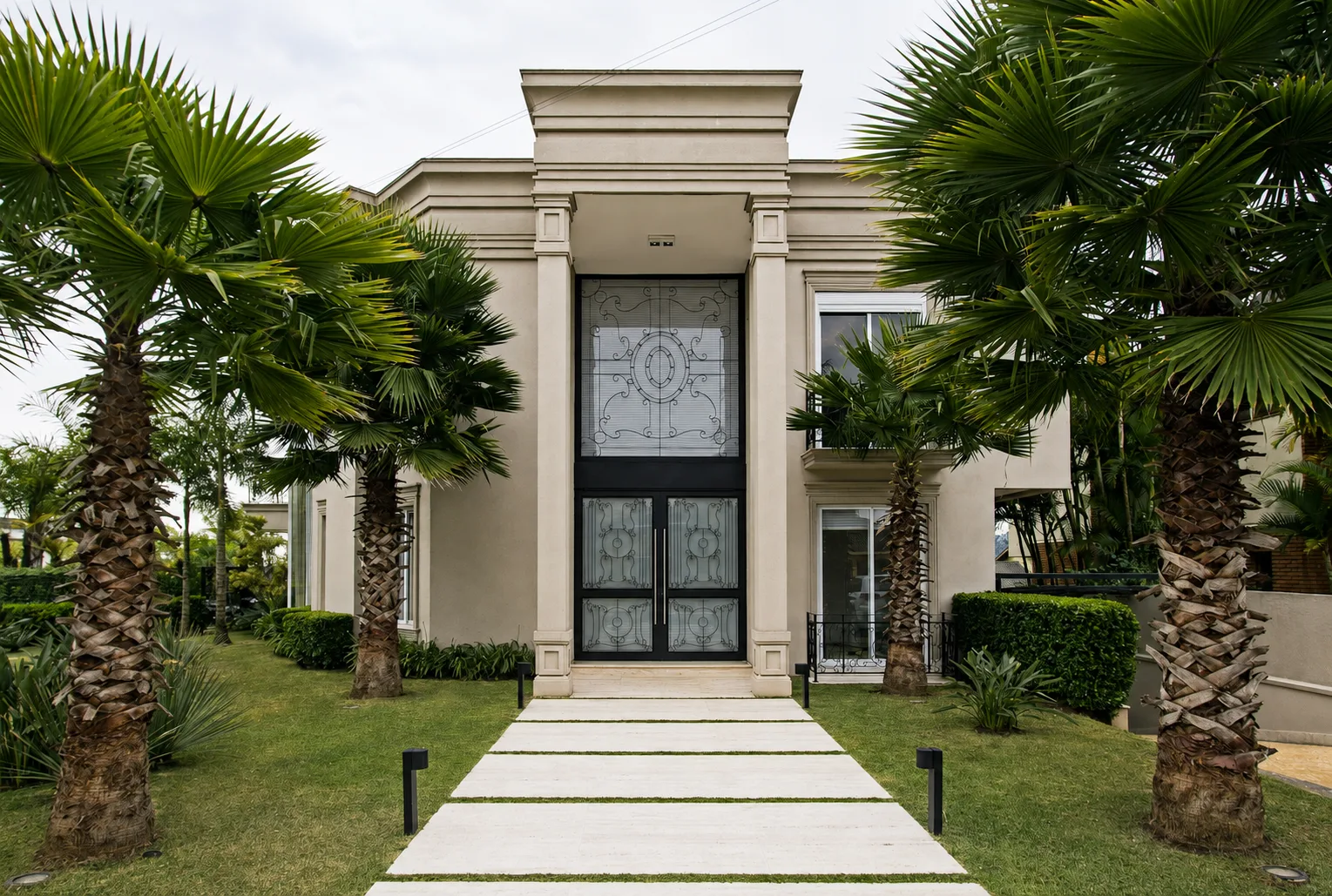 Fachada clássica de residência de alto padrão, com entrada monumental, volumes simétricos, grandes painéis de vidro, detalhes arquitetônicos elegantes e paisagismo com palmeiras.